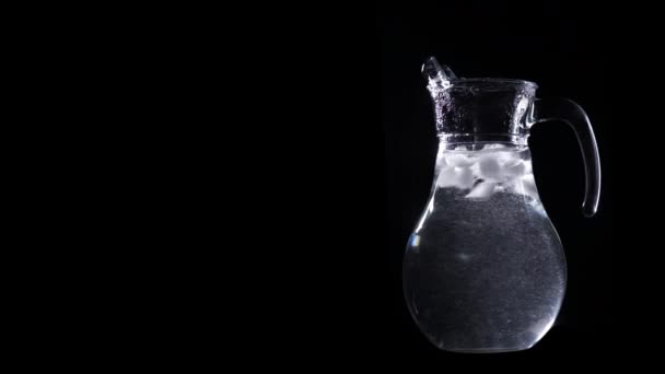 Concetto di acqua pulita. Acqua con ghiaccio ruotare in una brocca trasparente isolata su sfondo nero con spazio di copia per il testo a sinistra
. - Filmati, video