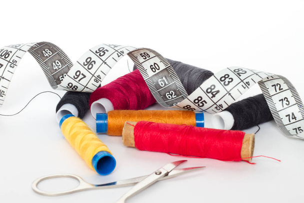 A spool of colored thread, an inch and scissors on a white background. Spool yellow, red, orange, gray and black threads, lying next to a centimeter and a pair of scissors. - Photo, image