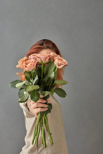 young red-haired woman holding bouquet of cappuccino roses against grey background and covering face, mother's day concept - Φωτογραφία, εικόνα