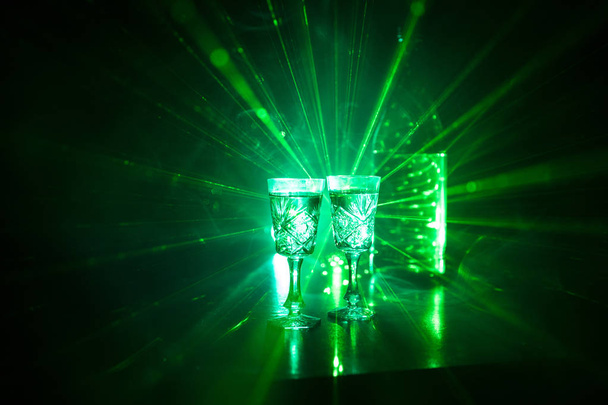 zwei Gläser Wodka mit Flasche auf dunklem, nebeligen Hintergrund mit leuchtenden Lichtern (Laser, Herd) mehrfarbig. Club Drinks Thema Dekoration. Leere Räume - Foto, Bild