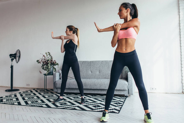 Two fit young woman stretching their arms exercising at home. Caucasian female fitness models working out in the morning. - Photo, Image