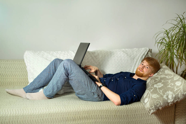 handsome young guy with a beard working at home with a laptop lying on the couch - Фото, изображение