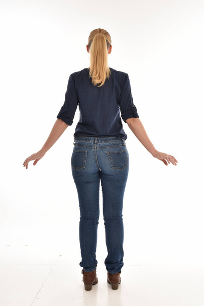 full length portrait of blonde girl wearing simple blue shirt and jeans, standing pose facing away from the camera. isolated on white background. - Fotografie, Obrázek
