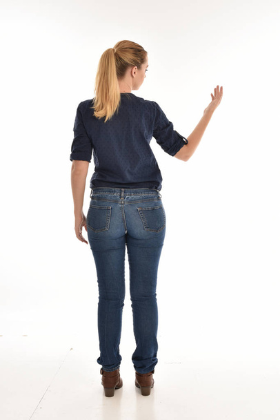 カメラから離れて直面しているポーズに立っているシンプルなブルーのシャツとジーンズを身に着けている金髪の女の子の完全な長さの肖像画。白い背景に分離. - 写真・画像