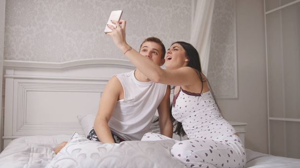 ベッド、魅力的な若い男、パジャマ姿で座っている女の子の selfie を作る幸せな陽気な愛情のあるカップル - 写真・画像