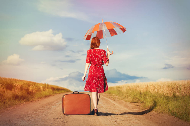Молодая рыжая девушка с зонтиком и сумкой идет по дороге в сельской местности. Путешествия по миру
 - Фото, изображение
