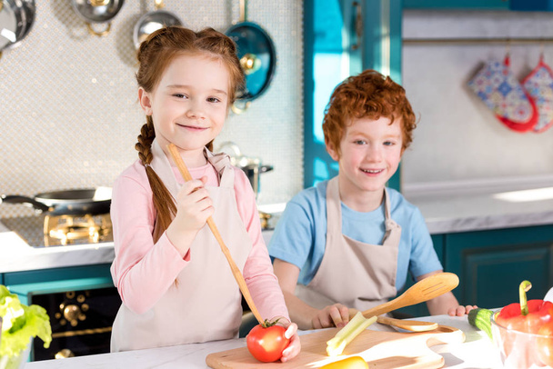 Urocza szczęśliwe dzieci w fartuchy uśmiechając się aparatu podczas gotowania razem w kuchni - Zdjęcie, obraz