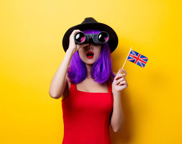 Portrait of young style hipster girl with purple hairstyle with binoculars and Great Britain flag in hand on yellow background - Photo, Image