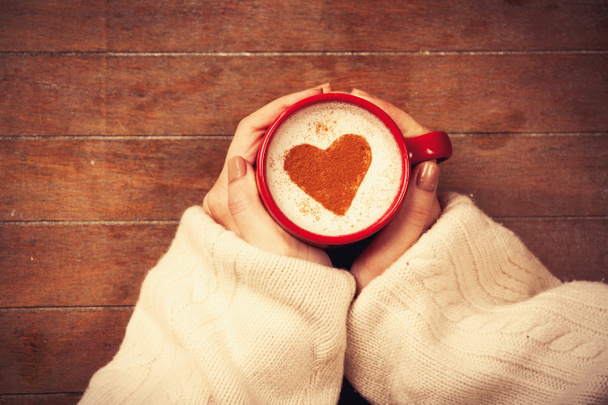 Female hand holding cup of cappuccino with heart shape cinnamon on wooden background - Photo, Image