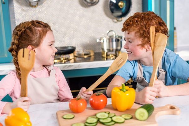 happy kids with wooden utensils smiling each other while cooking together in kitchen - Foto, afbeelding