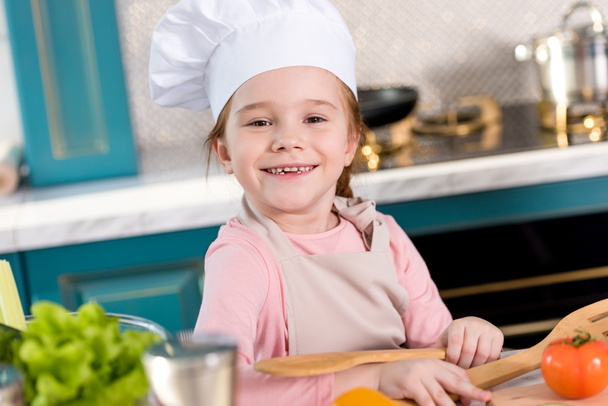 adorable niño en sombrero de chef y delantal sonriendo a la cámara mientras cocina en la cocina
 - Foto, imagen