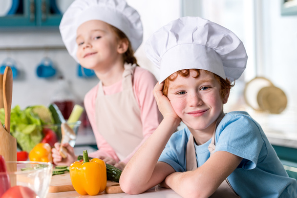 Urocza rodzeństwo w czapki szefa kuchni i fartuchy uśmiechając się aparatu podczas gotowania w kuchni - Zdjęcie, obraz