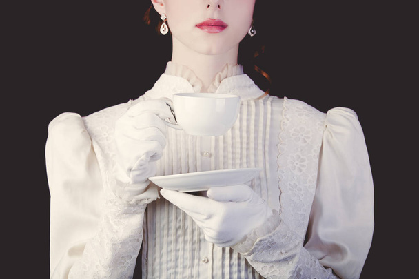 Young redhead woman in white victorian era clothes with cup of tea on black background. - Photo, Image