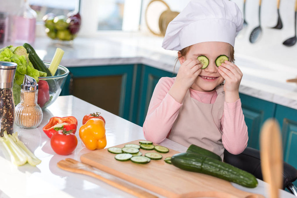 smiling child in chef hat and apron holding slices of cucumber while cooking in kitchen - Fotó, kép