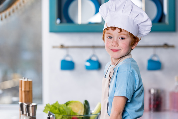 lindo niño en sombrero de chef y delantal sonriendo a la cámara en la cocina
 - Foto, imagen