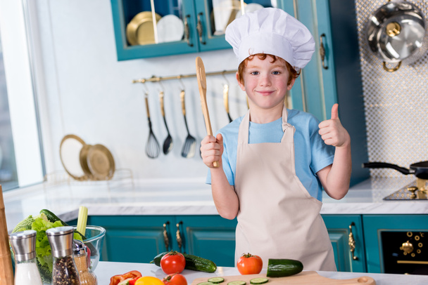 cute little boy in chef hat and apron smiling at camera and showing thumb up in kitchen - Foto, Imagen