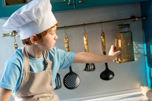 adorable niño en sombrero de chef y delantal mirando los utensilios mientras está sentado en la cocina
 - Foto, Imagen