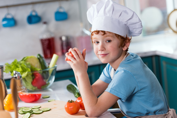 cute little boy in chef hat smiling at camera while cooking in kitchen - Valokuva, kuva