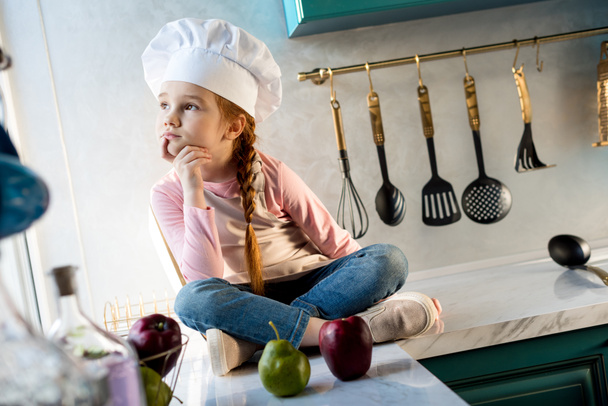pensive kid in chef hat looking away while sitting in kitchen - Fotó, kép