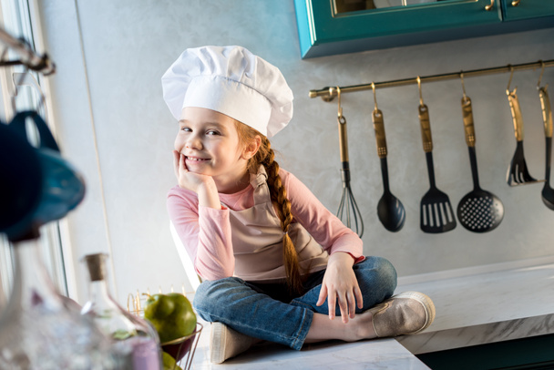 adorable child in chef hat sitting in kitchen and smiling at camera - Foto, immagini