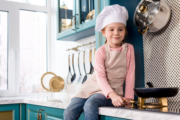 Cute usmívající se dítě v kuchař klobouk a zástěru, při pohledu na pánev na sporák - Fotografie, Obrázek
