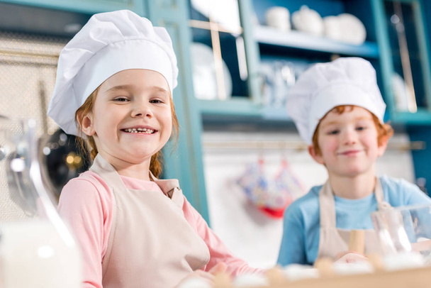 adorable children in chef hats and aprons smiling at camera while cooking together in kitchen  - Fotografie, Obrázek