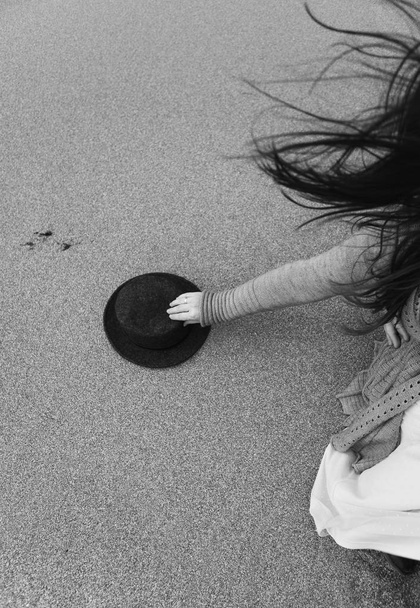 the girl's hand reaches for her hat, her dark hair fluttering in the wind, she sits on the sand - Photo, Image