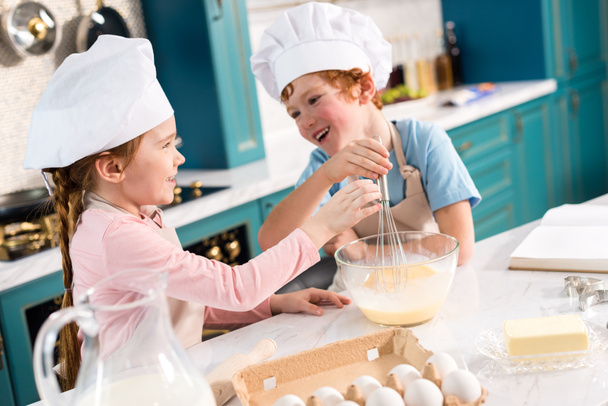cute little kids in chef hats smiling each other while whisking dough in kitchen - Foto, immagini