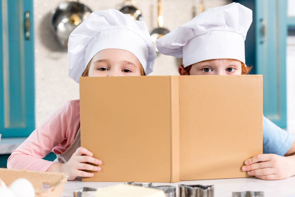 niños lindos en sombreros de chef sosteniendo libro de cocina y mirando a la cámara
  - Foto, imagen