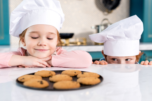 szakács sapka nézett finom sütiket a táblázat-imádnivaló boldog gyerekek - Fotó, kép