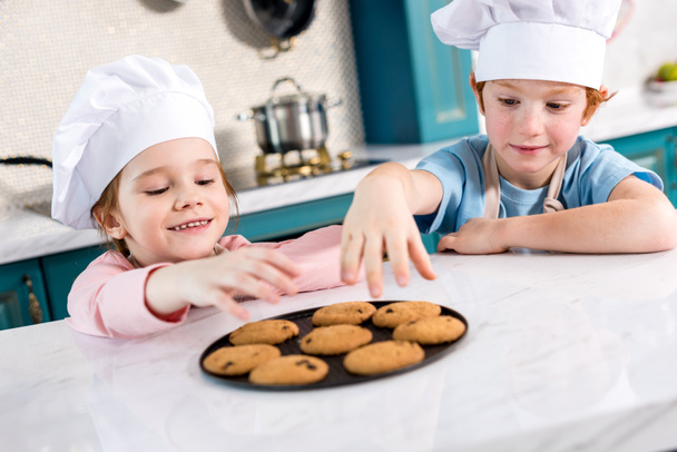 happy little kids in chef hats eating tasty cookies in kitchen - Fotó, kép
