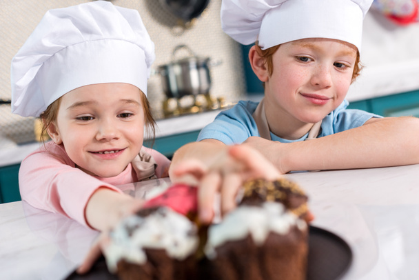 smiling little children in chef hats looking at delicious cupcakes on foreground  - Foto, imagen
