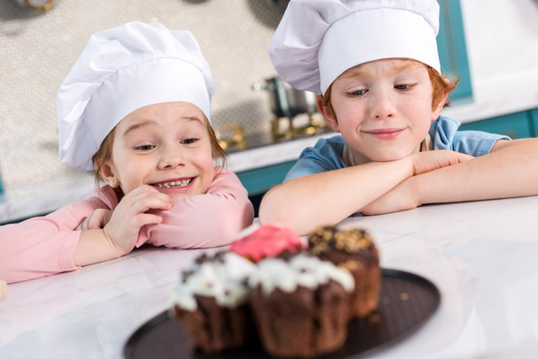 szczęśliwe dzieci w kapeluszach szefa kuchni, patrząc na pyszne babeczki na pierwszym planie  - Zdjęcie, obraz