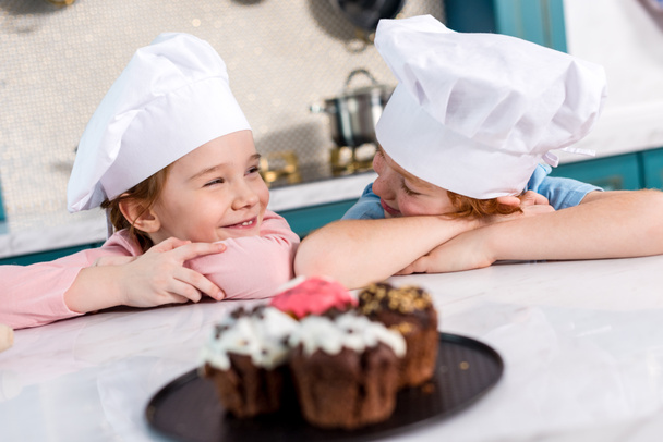 adorable kids in chef hats smiling each other and delicious cupcakes on foreground - Фото, зображення