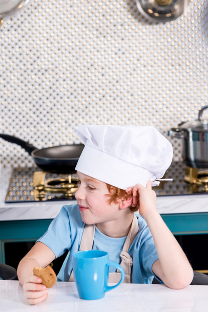 cute little boy in chef hat drinking tea and eating cookie in kitchen - Фото, изображение