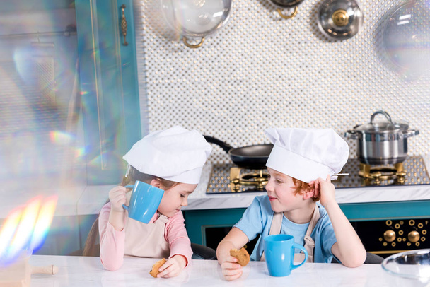 schattig weinig kinderen in chef-kok hoeden drinken thee en koekjes eten in de keuken - Foto, afbeelding