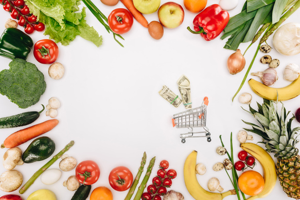 bovenaanzicht van kleine winkelwagen met dollars tussen groenten en fruit geïsoleerd op wit - Foto, afbeelding