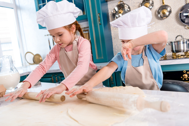 сосредоточенные маленькие дети в шляпах шеф-повара и фартуках катящихся тесто на кухне
 - Фото, изображение