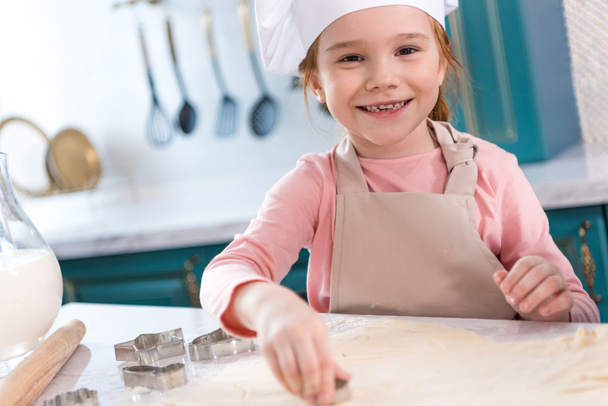 adorable child in chef hat and apron smiling at camera while preparing cookies - Fotoğraf, Görsel