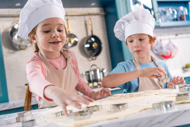 urocze małe dzieci w kapeluszach szefa kuchni i fartuchy razem przygotowanie plików cookie - Zdjęcie, obraz