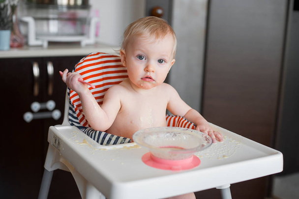 Little cute girl sitting in high chair and learning to eat with spoon herself. Funny happy baby smudge food on the table - Foto, Imagem