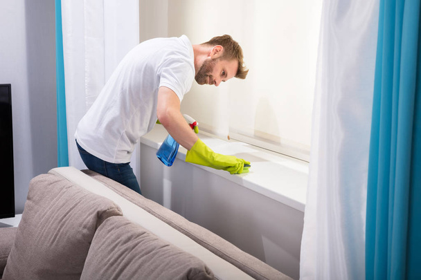 Young Male Janitor Cleaning Windowsill With Sponge In Living Room - Foto, immagini