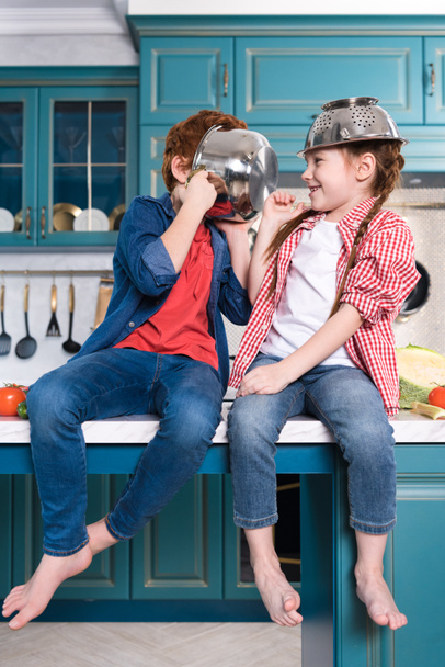 αξιολάτρευτο ξυπόλητα παιδιά διασκεδάζουν με οικιακά σκεύη, ενώ κάθεται στο τραπέζι της κουζίνας  - Φωτογραφία, εικόνα