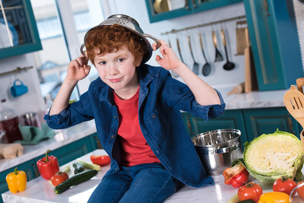cute little boy with pan on head smiling at camera while sitting on kitchen table  - Foto, Bild