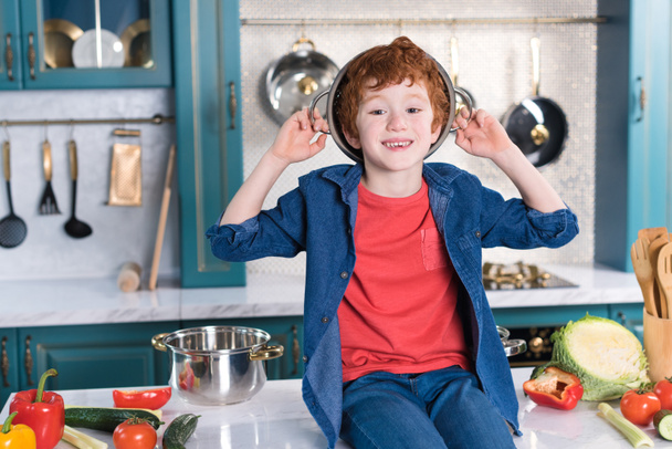 αξιολάτρευτο μικρό αγόρι με τηγάνι στο κεφάλι χαμογελώντας σε κάμερα ενώ κάθεται στο τραπέζι της κουζίνας  - Φωτογραφία, εικόνα
