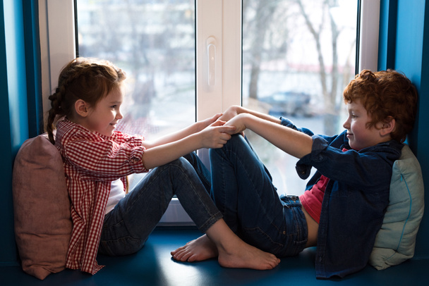 adorable little kids holding hands and smiling each other while sitting on windowsill - Fotoğraf, Görsel