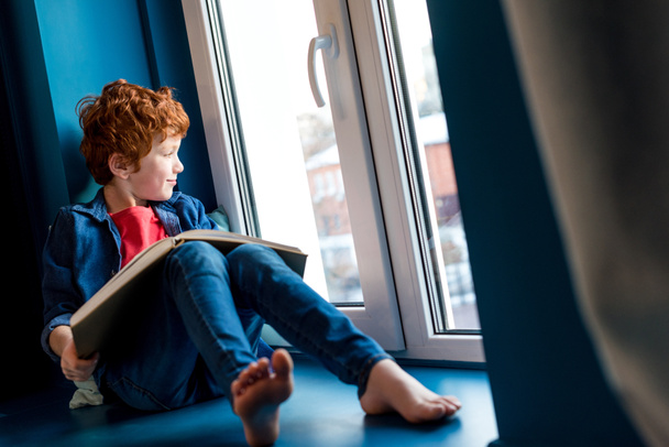 cute barefoot boy with book sitting on windowsill - Zdjęcie, obraz