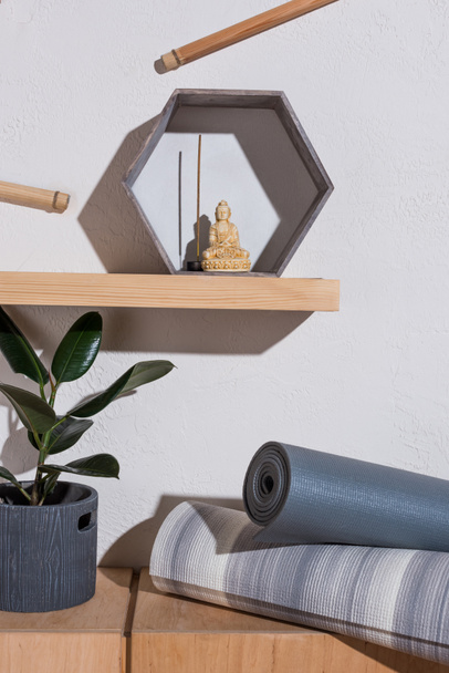 small statue of buddha in frame with incense stick on wooden shelf, yoga mats near plant - Photo, Image