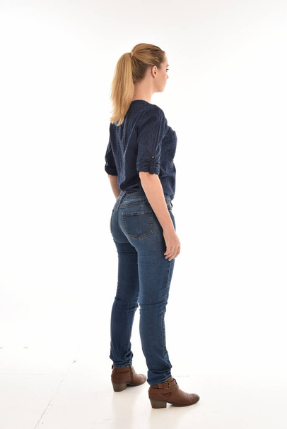 Portret van de volledige lengte van blond meisje dragen van eenvoudige blauw shirt en spijkerbroek, staande pose geconfronteerd weg van de camera. geïsoleerd op witte achtergrond. - Foto, afbeelding