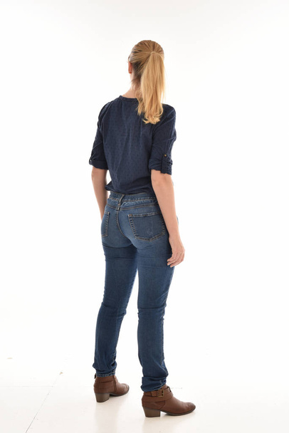 full length portrait of blonde girl wearing simple blue shirt and jeans, standing pose facing away from the camera. isolated on white background. - Fotografie, Obrázek